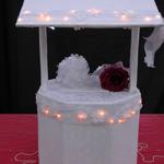 White Lighted Wishing Well: White organza fabric w/ flowers, over white satin. Organza ribbon trimmed w/ flowers circles the well & the roof of well. Soft lights illuminate from behind the ribbon. Bow accents cover of well, trimmed with pearls.  Gentle spotlight illuminates bow. Very romantic.
